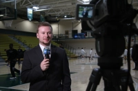A sports journalist student stands in front of a camera while holding a microphone. He stands in a gymnasium while basketball players are playing behind him.